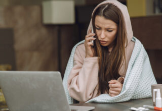 A sick woman working from home, wrapped in a blanket and wearing a hoodie, talks on the phone while sitting at a desk with a laptop, medicine, and a cup—possibly calling her doctor about flu or cold symptoms.