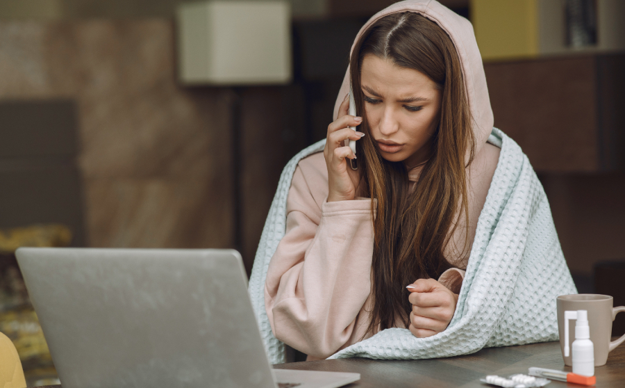 A sick woman working from home, wrapped in a blanket and wearing a hoodie, talks on the phone while sitting at a desk with a laptop, medicine, and a cup—possibly calling her doctor about flu or cold symptoms.