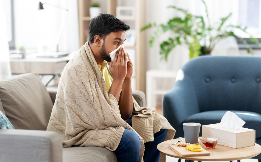 Man on sofa blowing nose with tissue. sicknote.com. He appears unwell